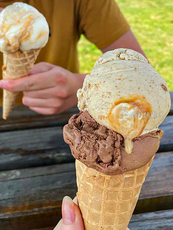 特写照片上的人拿着华夫饼，冰淇淋甜筒里有两勺巧克力和咸味焦糖冰淇淋，一对夫妇坐在公园长凳上，阳光灿烂，聚焦前景