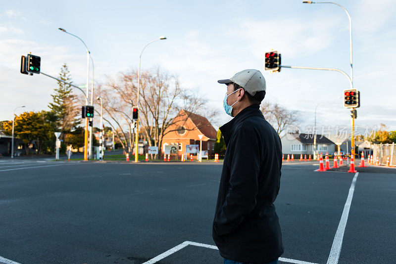 戴着口罩的男子在十字路口等待绿灯。沿街的道路交通锥。