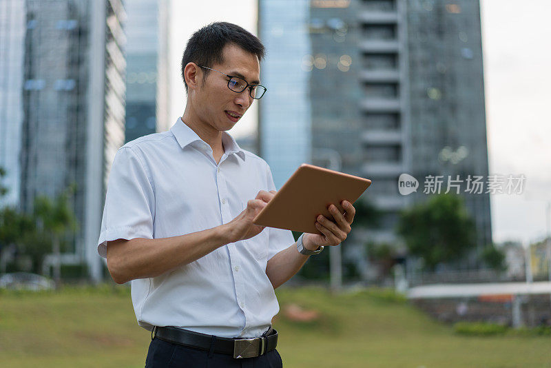 一位亚洲男性白领正在办公大楼外的草地上使用平板电脑