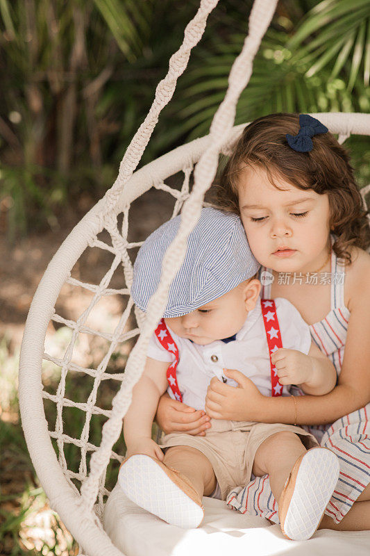 在南佛罗里达7月4日的庆祝活动中，一个6个月大的小男孩穿着红色背带和蓝色领结，他3岁的大姐姐抱着他，穿着红白蓝条纹的裙子，他们在波西奥悬挂的椅子上荡秋千