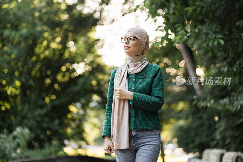 欢快的年轻伊斯兰妇女与头巾走在城市夏季公园。