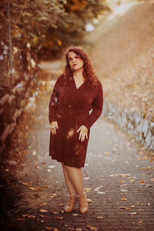 年轻优雅的红发女子肖像穿着酒红色连衣裙，在秋季公园摆姿势