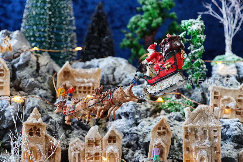 特写图像的圣诞老人雪橇和驯鹿飞过风景优美的圣诞村显示，人工塑料和木制圣诞树在山上设置，照明自制木屋，重点在前景