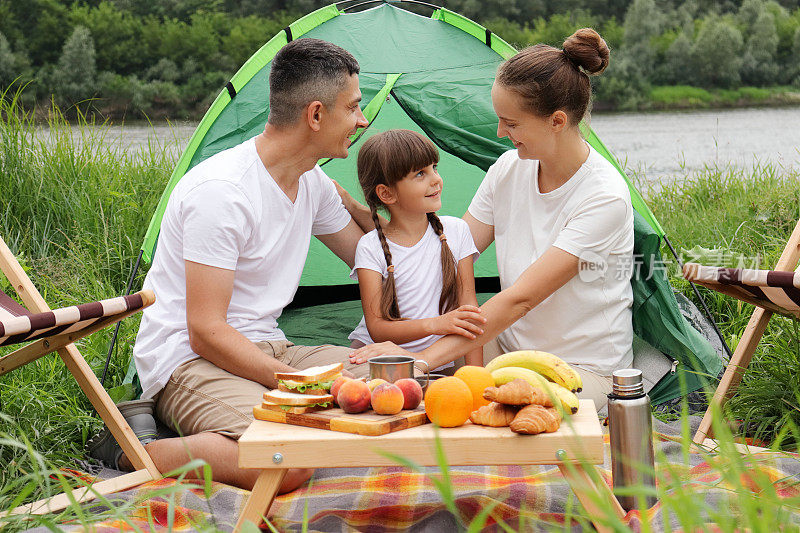 家人在河边休息。在自然公园的河边露营，夫妇和女儿坐在帐篷旁。周末在户外。