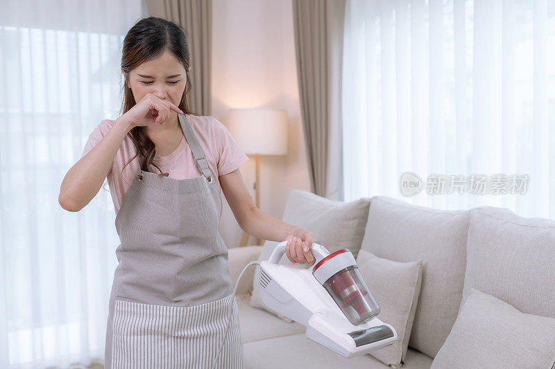 年轻的亚洲女子在假期的空闲时间打扫家里的客厅，她鼻塞，打喷嚏，因为对灰尘过敏。