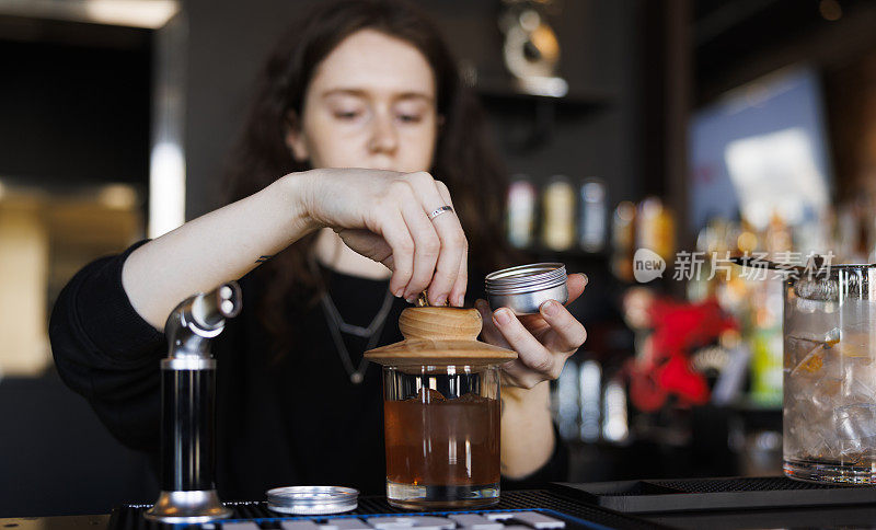 女调酒师在酒吧里调制熏制老式鸡尾酒，用木盖盖住玻璃杯，在吧台数帽子。把注意力放在饮料和手上，特写。