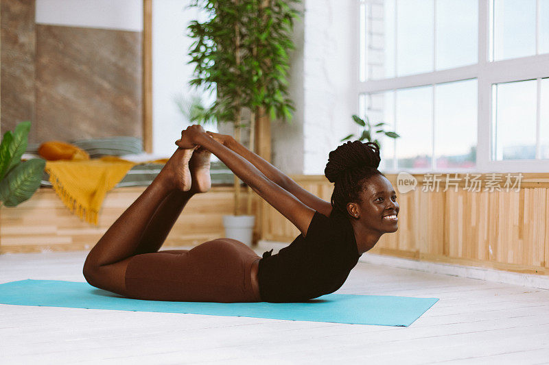 美丽和运动的非洲妇女躺在弓姿势侧视图微笑，做瑜伽的体操垫上装饰的工作室。肌肉伸展，体操练习，健身，保持身体健康