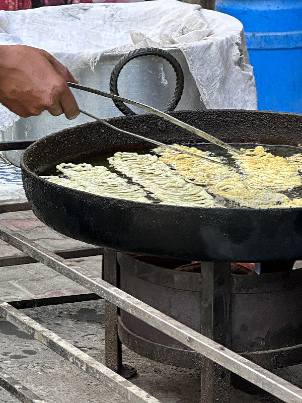 贾勒比油炸面糊甜点的图片，在户外炉子上制作的街头食品，厨师用钳子把金色的螺旋面糊翻动，在煎锅里煮油，重点放在前景