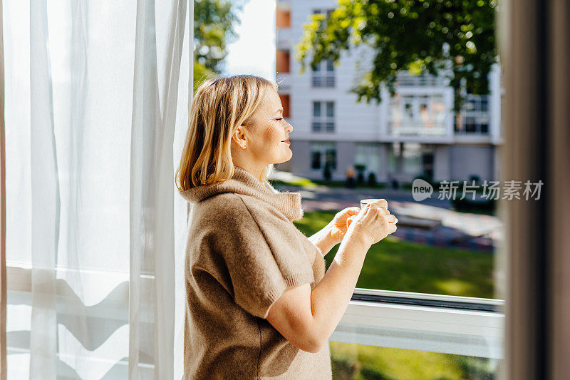 自由放松的时间。美丽的女人坐在家里白色的窗台上，读着纸质书，端着杯子，喝着热咖啡。快乐的女士拿着杯子在窗口边休息，复印空间