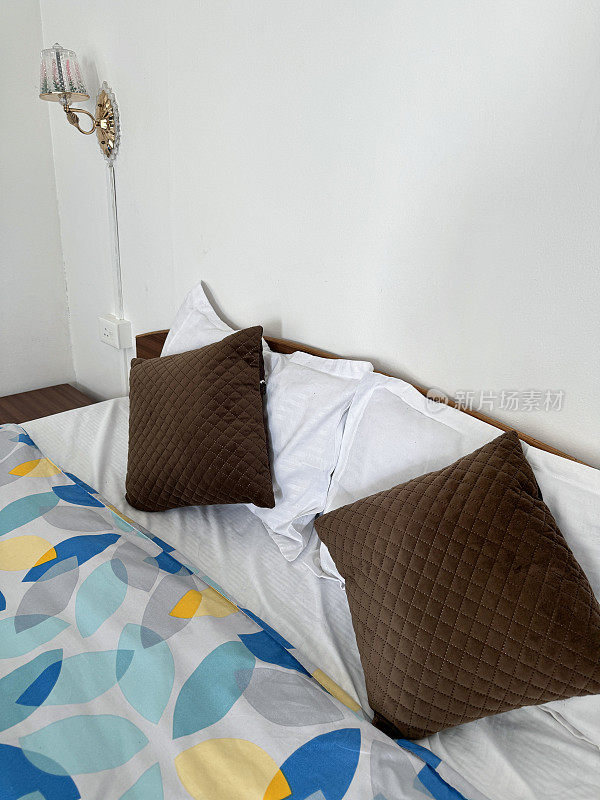 木制床头板的双人床的特写图像，复古风格的棕色绗缝靠垫堆叠在白色枕头上，图案被套，木制床头柜上的玻璃罩壁挂灯，重点放在前景上，抬高视野