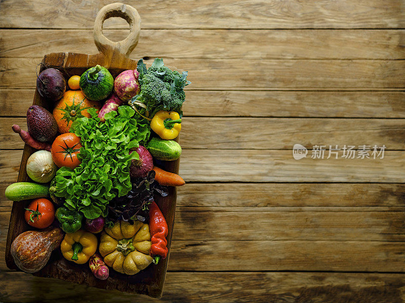 许多色彩鲜艳的健康蔬菜陈列在一个古色古香的实木木雕碗上，在一个老柚木镶板的桌子背景上，阴影大气的乡村情绪，与良好的复制空间在右侧的形象。