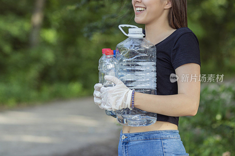 女志愿者穿着衣服在公园里捡垃圾和塑料垃圾。小女孩戴着手套，在户外把垃圾放进黑色的塑料袋里。