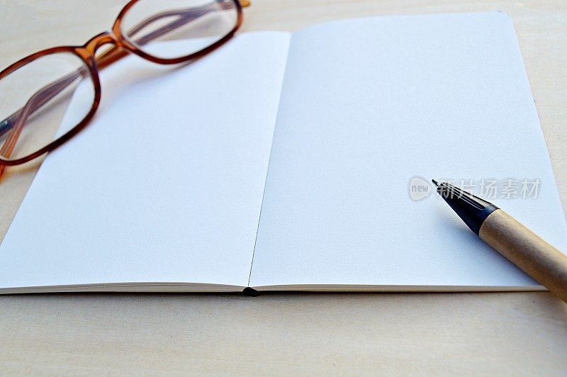 一张水平的照片，白色的空白页的记事本，连同一支棕色和黑色的钢笔和一副眼镜，放在一个木制的米色水平背景上，很有美感。