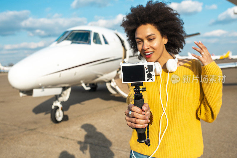 一名年轻的视频博主在机场停机坪上的一架私人飞机旁用自拍杆上的相机拍摄自己