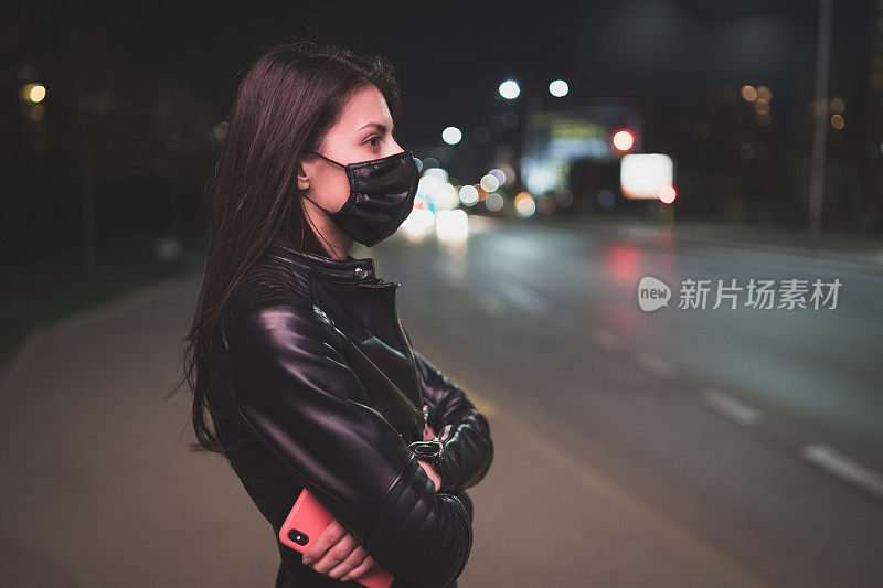 深色头发的女人，穿着黑色皮夹克，戴着黑色面罩，在晚上下班后等着公交车