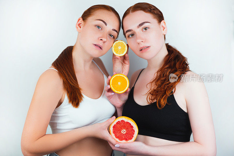 两个红头发的年轻女子手里拿着一半的葡萄柚和两半的橙子站在孤立的白色背景，美丽的肖像概念