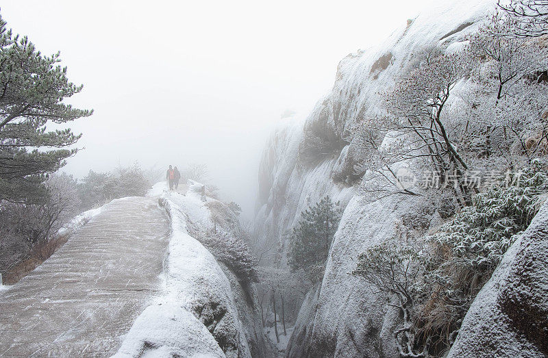 两个旅行者走在人行道上，冬天，黄山上到处都是积雪，天气很冷。它是联合国教科文组织世界遗产之一。