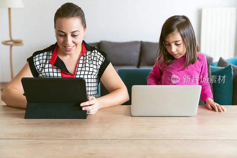 商业女性在家里使用数字平板电脑工作，同时照顾看着笔记本电脑的女儿