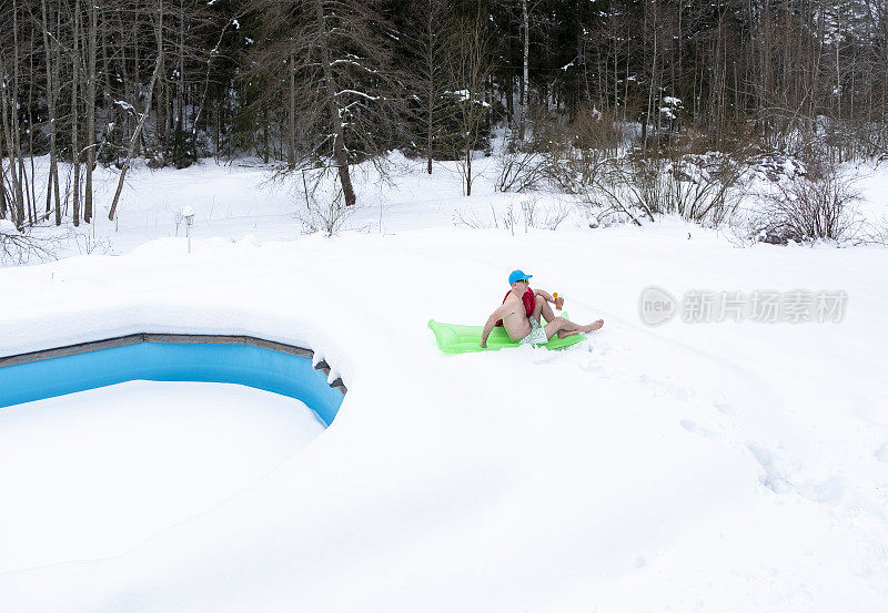 一个男人穿着泳裤，戴着墨镜，在一个充气床垫上结冰。冬天，一个男人在房子后院里的池塘边的雪地里休息。