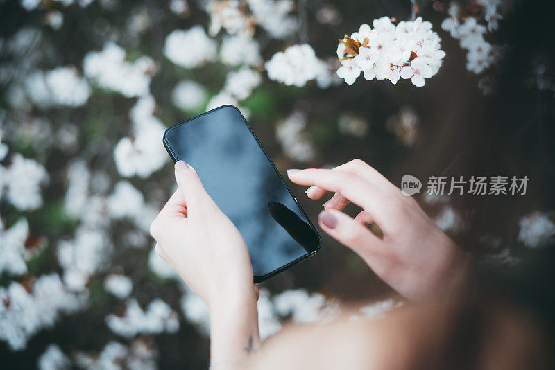 在一个樱花盛开的公园里，一名女子在用智能手机拍照