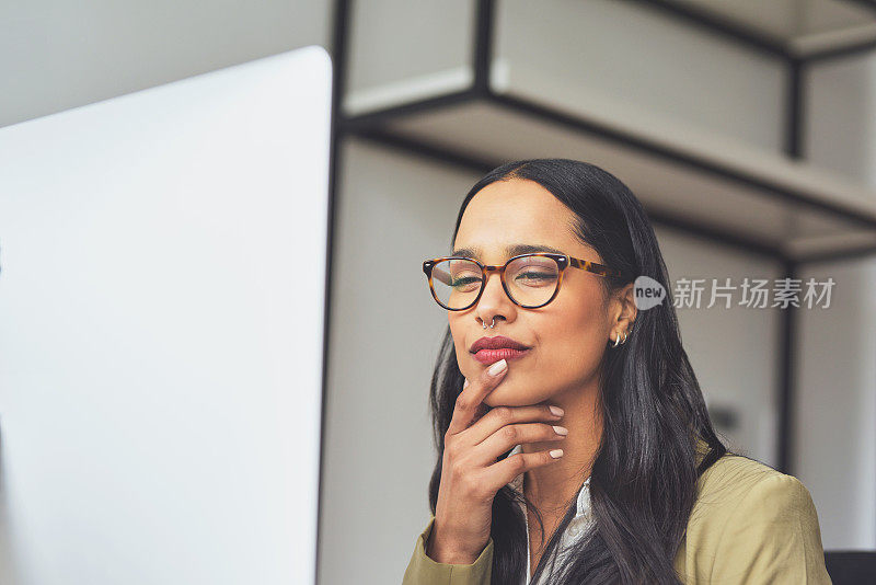 一个迷人的年轻女商人独自坐在她的办公室里，看着沉思，同时使用她的电脑