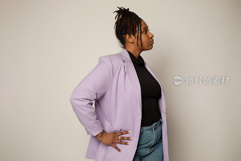 非裔美国女性，穿着牛仔裤和淡紫色运动夹克，手放在臀部，目光移开