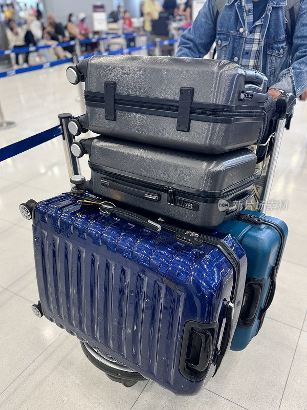特写照片中，无法辨认的乘客穿着牛仔夹克，操纵着一堆轮式行李箱的行李车，在支柱和可伸缩皮带旁排队，机场出发区，闪亮的瓷砖地板，重点放在前景上