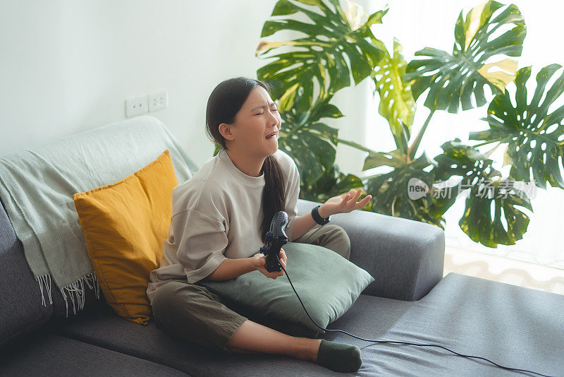 一名亚洲女子在输了游戏后坐在家里的沙发上，用操纵杆玩游戏，感到很沮丧。