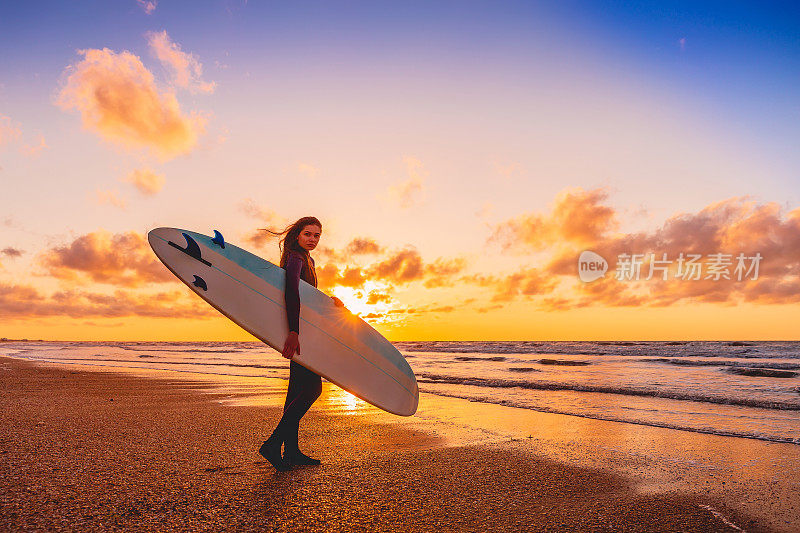 冲浪女孩带着长板去冲浪。日落时分，一个女人在海滩上带着冲浪板。