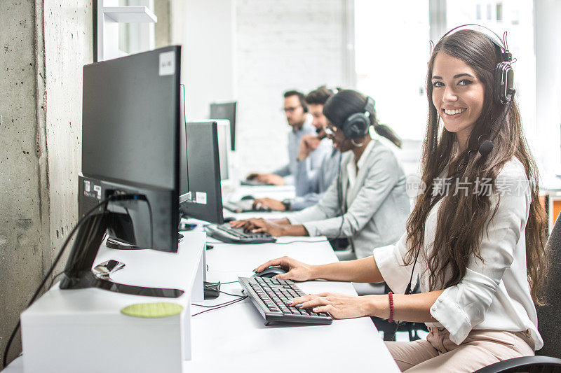 美丽微笑的女性客户支持工作人员与耳机使用计算机在呼叫中心。