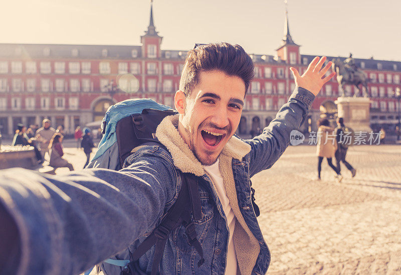 在西班牙马德里市长广场，一名年轻的旅游学生背着背包，兴致勃勃地用智能手机自拍一张笑脸。度假、环游世界的概念。