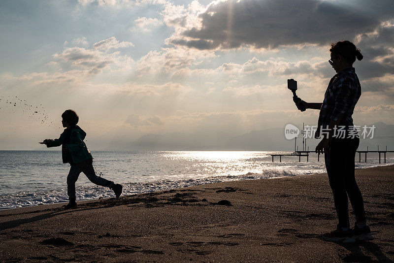 年轻女子用智能手机在海滩上拍摄男孩