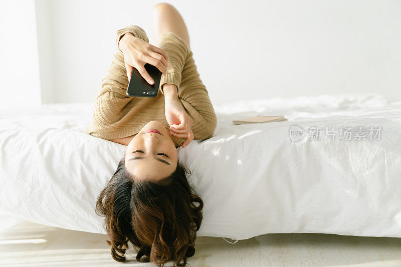 美丽的年轻亚洲妇女与她的头发下垂的边缘躺在她的背部，她的膝盖微微抬起，在床上使用手机在卧室的冬天。休息放松的生活理念。