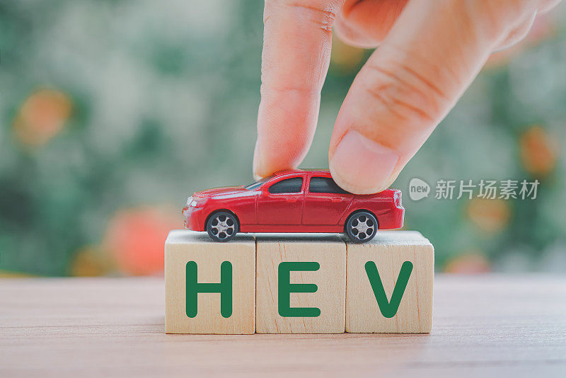 手持红色微型汽车的木制立方体上面带有HEV字样，在模糊的自然背景上意味着拯救世界的混合动力电动汽车运动，替代概念