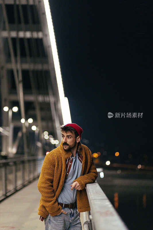 一个年轻英俊的时髦男人的肖像与胡子倚靠在桥栏杆在晚上