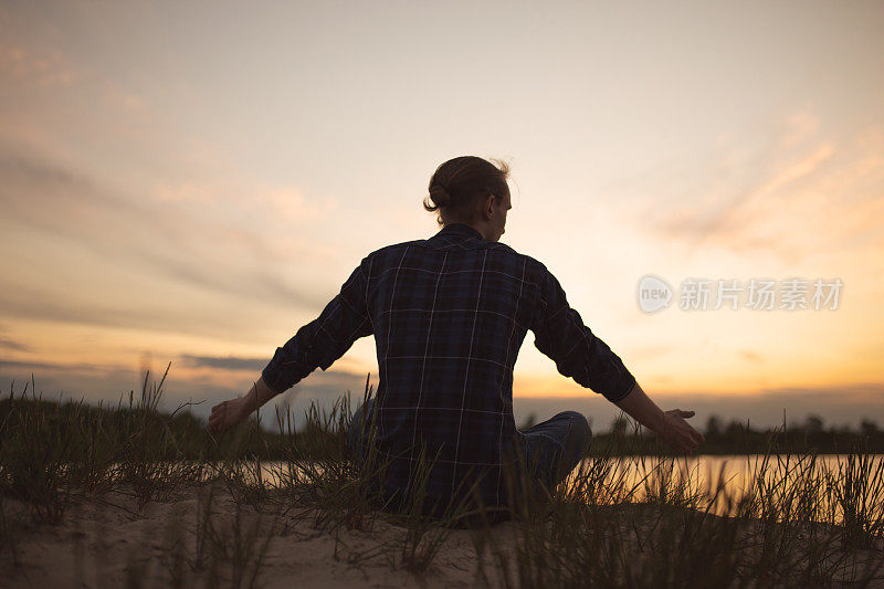 美丽的夏日夕阳背景上的男子剪影。橙色的天空。那家伙正坐在河边。人坐在河岸上