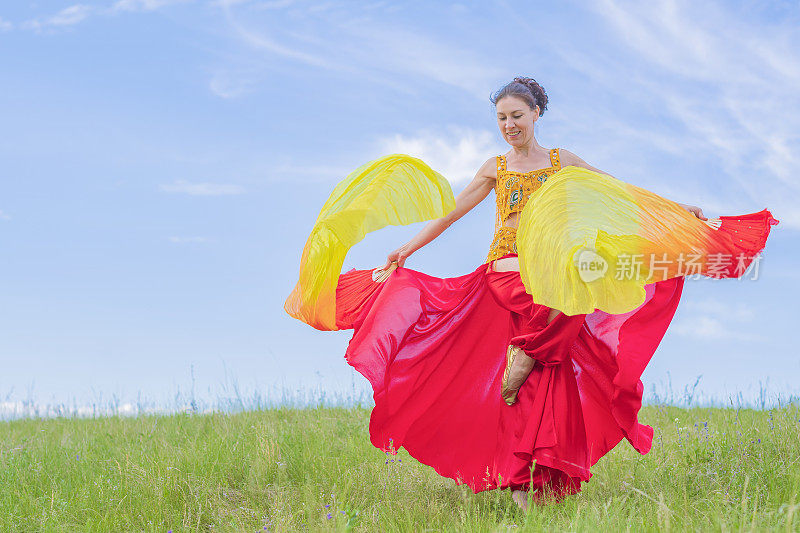 身着红色民族服装的美女在草地上的开阔天空下挥舞着扇子跳着东方舞蹈