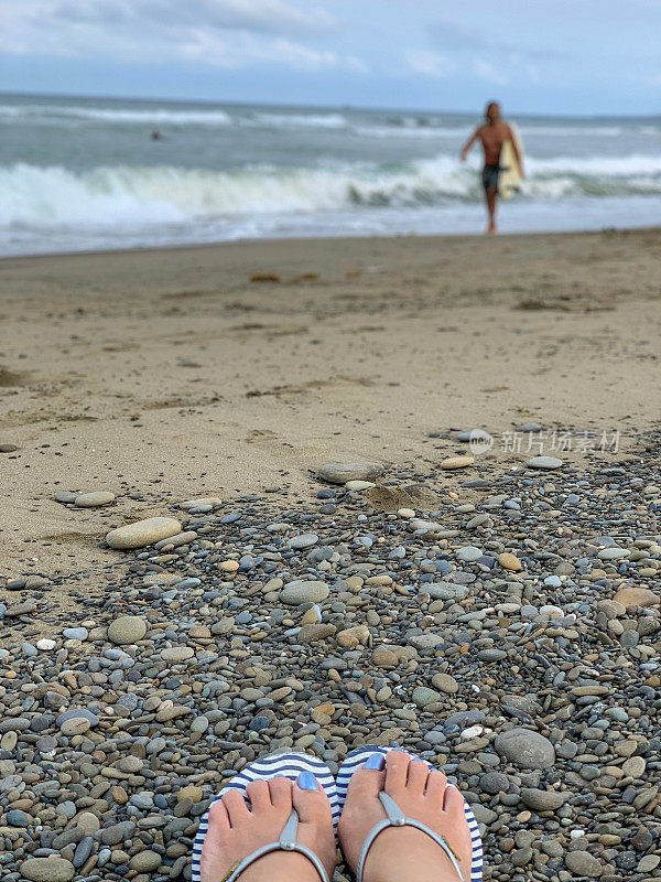 一个冲浪者完成了冲浪，拿着冲浪板在岸上休息，前景是一双穿着人字拖的女人的脚