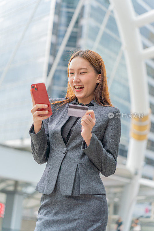 一名年轻的亚洲女商人穿着灰色的办公套装，在商业区用她的智能手机使用信用卡进行在线交易，她微笑着自信