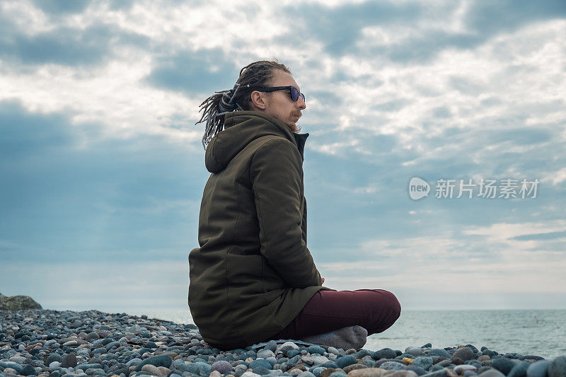 年轻男子与脏辫练习瑜伽冥想坐在海边的户外莲花位置。心理健康