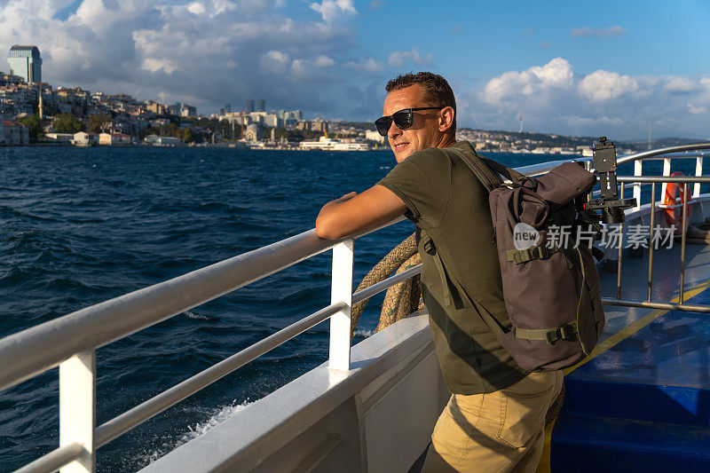 在一个阳光明媚的日子里，快乐的年轻男游客背着背包在伊斯坦布尔的渡轮上享受着大海和这座城市。旅游的概念，旅游
