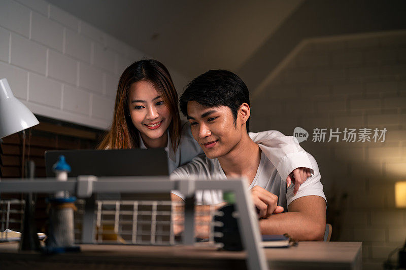 亚洲年轻人晚上在家和妻子在卧室里加班。迷人美丽的妻子穿着睡衣等着丈夫用笔记本电脑工作，在家里睡觉时上网交流。