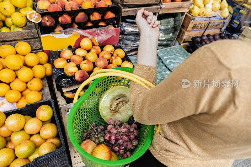 在街边的杂货店里，一位顾客举着购物篮挑选水果和蔬菜的特写