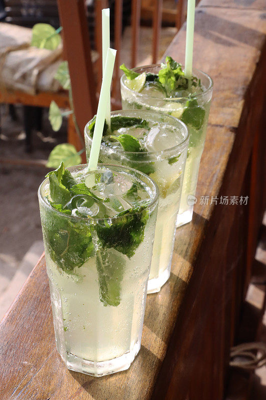 在露天海滩酒吧餐厅的木栏杆上，一排三杯鸡尾酒的图片，莫吉托鸡尾酒的玻璃杯，冰块，薄荷叶枝和酸橙片，吸管，沙子背景，高架视图，重点在前景