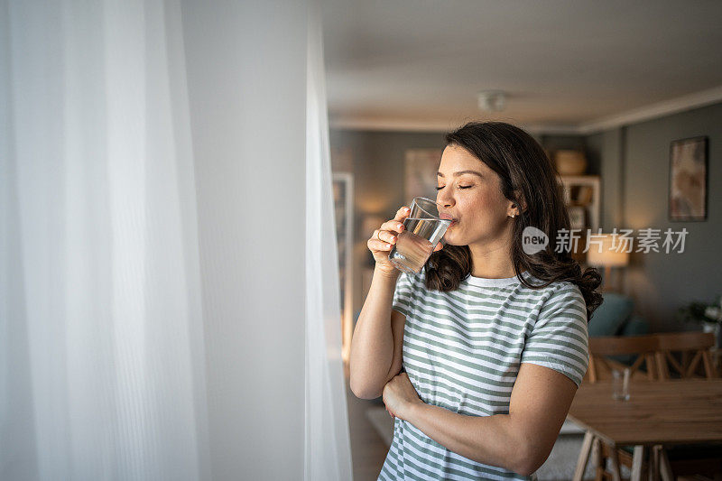 一个年轻女子站在窗边，用玻璃杯喝水。她的微笑反映了她关心自己的健康和适当摄入液体的意识。