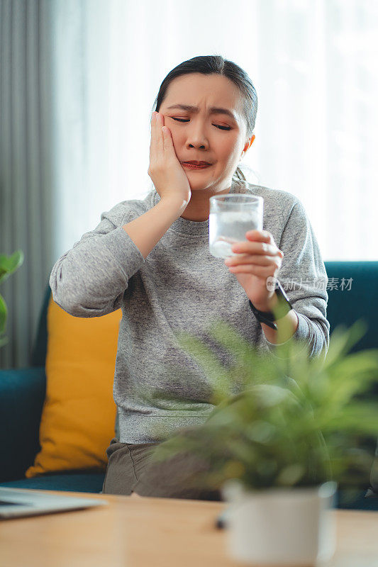 亚洲女性坐在客厅的沙发上，喝了凉水，摸了摸脸颊，就会感到牙齿敏感。