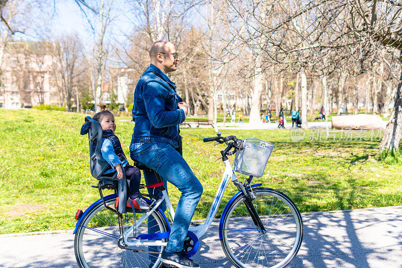 无忧无虑的爸爸和女儿在公园里骑车。慈爱的父母骑着自行车在公园里兜风，让他的孩子坐在儿童座椅上。