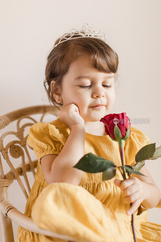 2岁的蹒跚学步的小女孩穿着黄色的连衣裙，戴着头冠，看着一朵红玫瑰