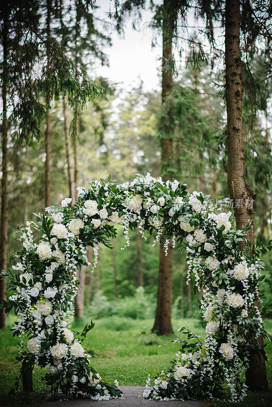 美丽的婚礼乡村装饰在森林。拱门上装饰着鲜花和绿色植物，后院是绿色植物。婚礼宴会厅。原来的花卉装饰。露天休息区。