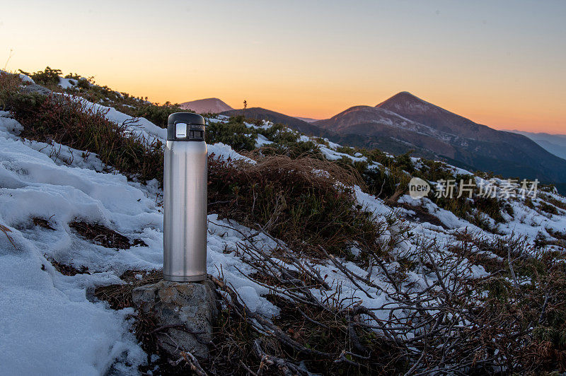 旅行用保温瓶装热饮。山顶上的热水瓶。热水瓶，雪，石头和山。外面有个保温杯。